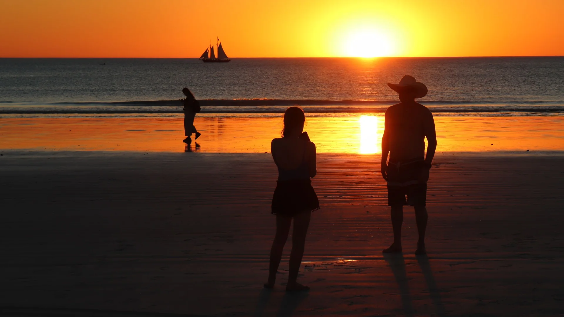 Two people stand in silhouette on the wet sand of Cable Beach as the sun sets over the Indian Ocean, with a sailing ship on the horizon