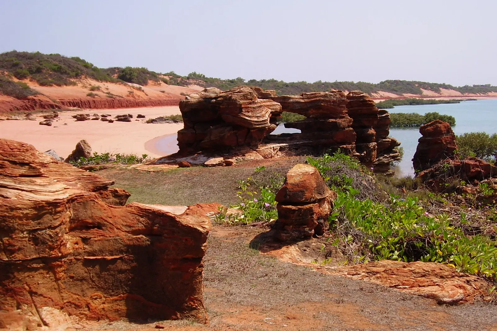 A pale sand beach in Roebuck Bay with red pindan rock stacks, scrub and a shallow bay behind